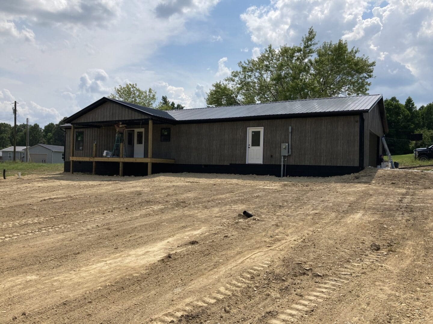 Newly constructed black wooden house with a porch on a dirt lot under a cloudy sky.